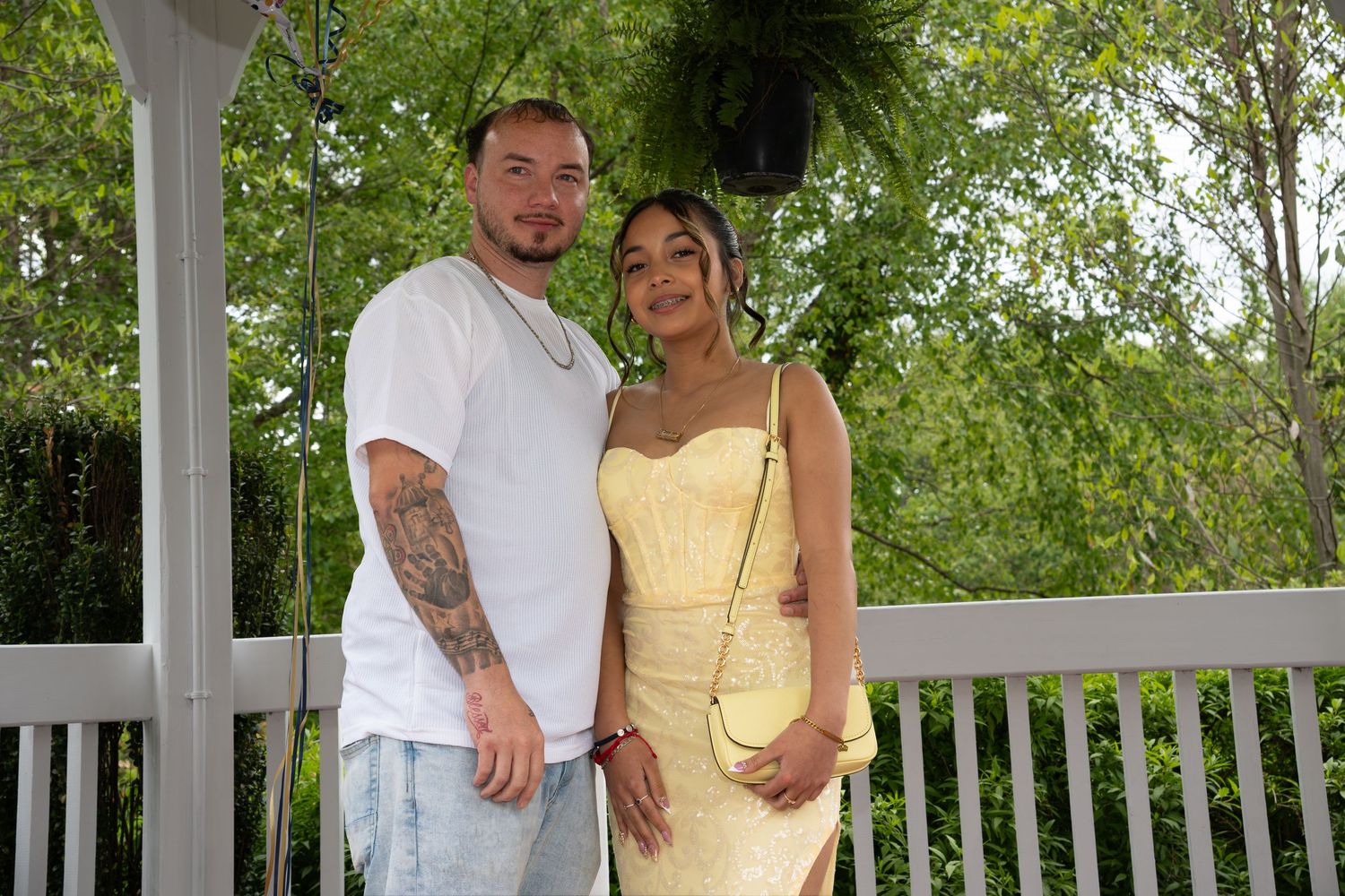 Smiling father and daughter posing on wooden deck, woman in yellow lace dress, man in white shirt with tattoos.