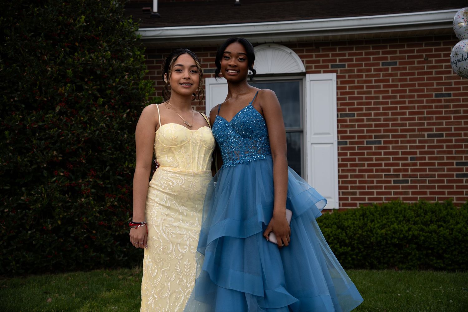 Two teenage girls in formal social dresses posing outdoors, one in yellow gown, one in blue tulle dress.