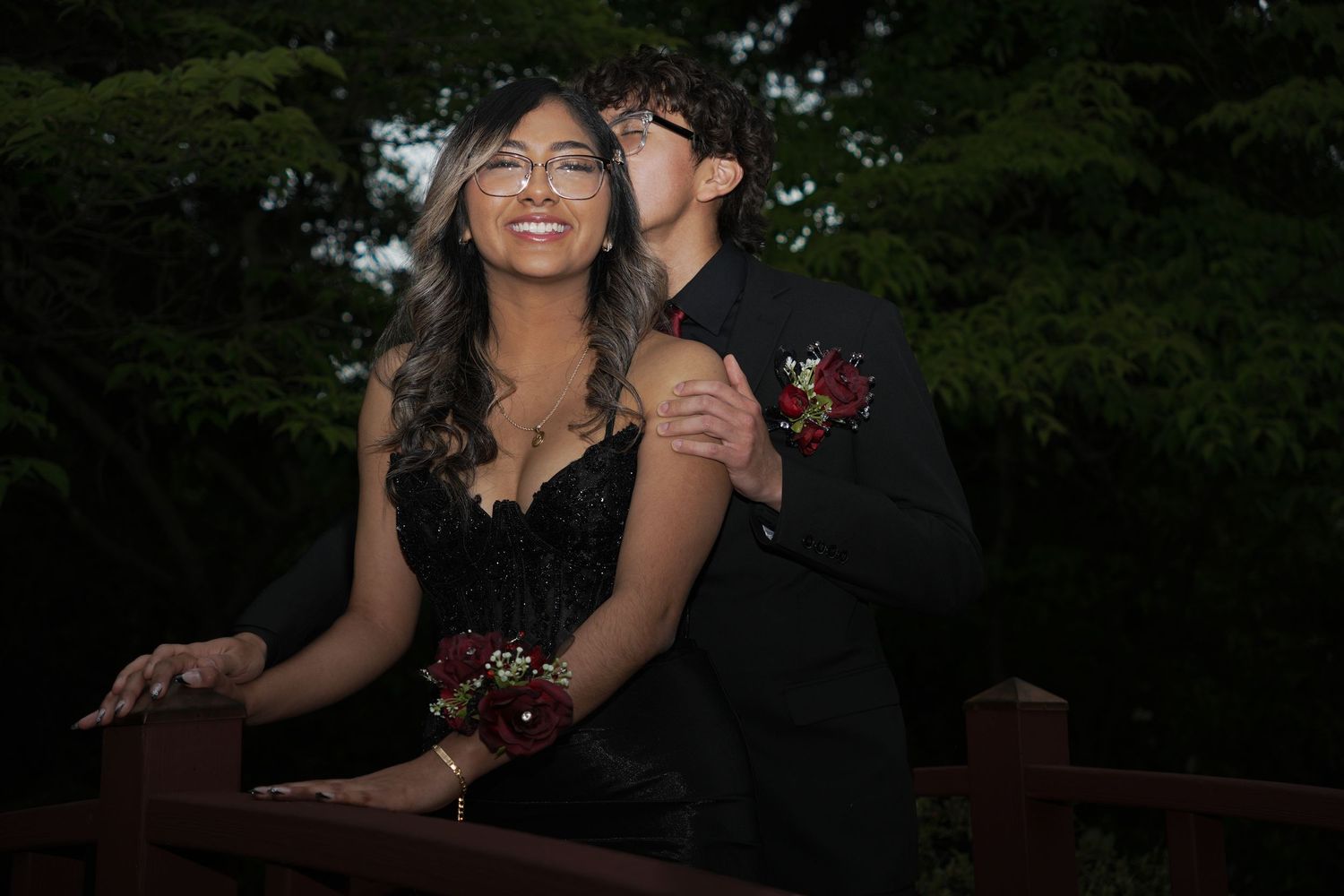 Couple in formal prom attire posing outdoors, girl in black dress with corsage, boy in suit with boutonniere kissing her cheek.