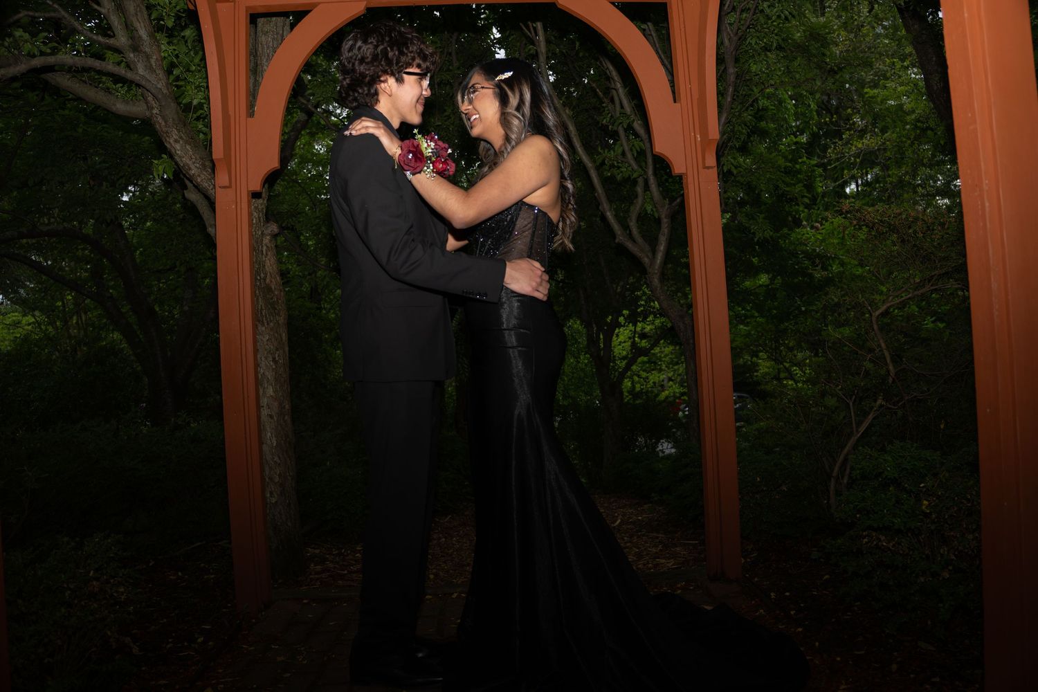 Couple in elegant black formal wear posing romantically under a wooden arch surrounded by lush green trees at night.