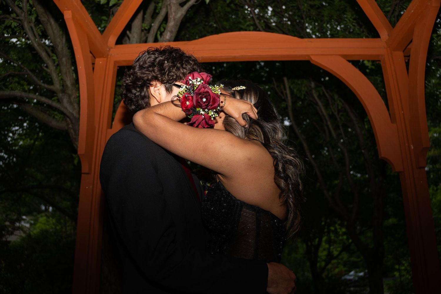 Couple sharing a romantic kiss under a wooden gazebo at night, bride holding a red bouquet.