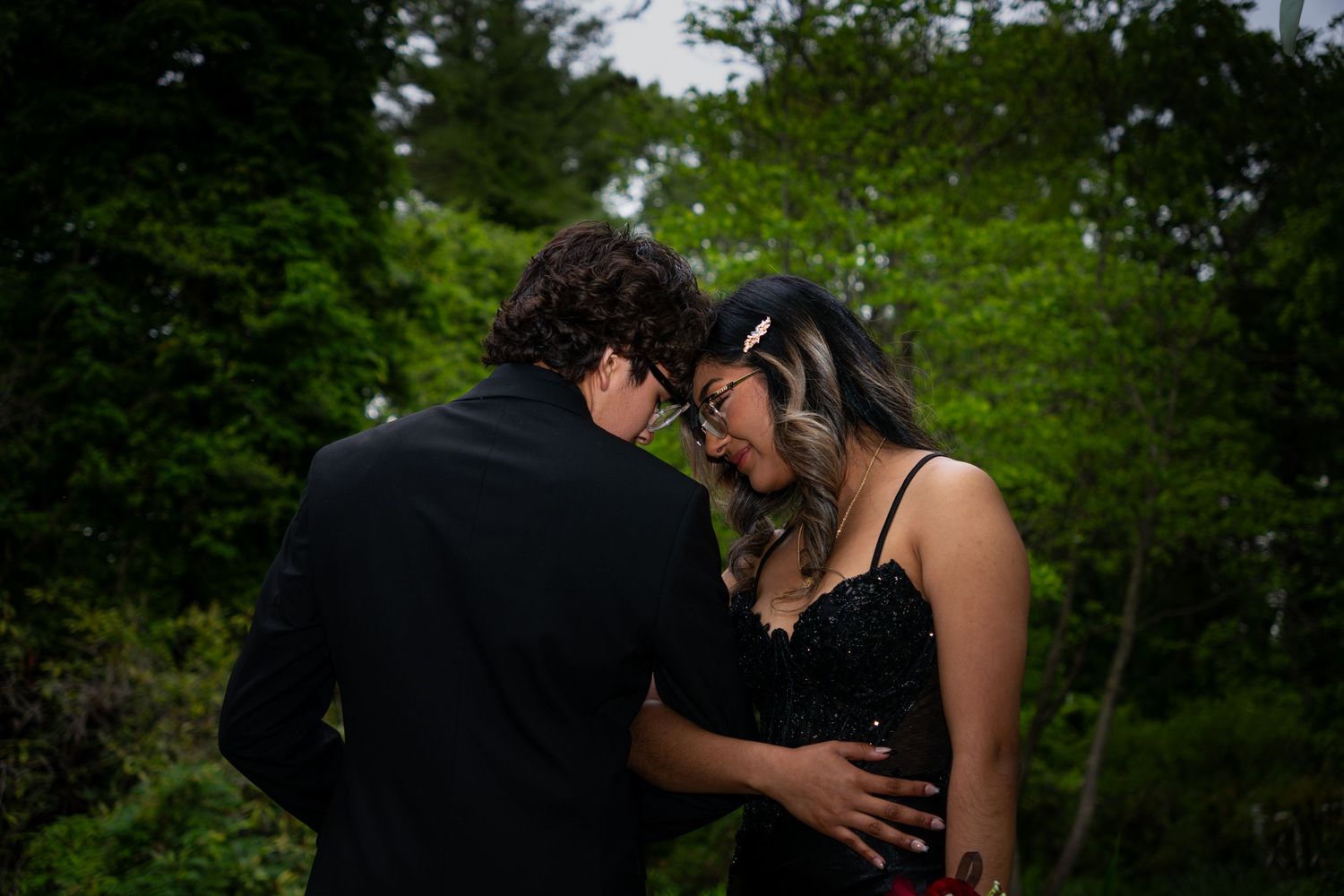 Couple in formal attire sharing an intimate moment surrounded by lush green trees outdoors.