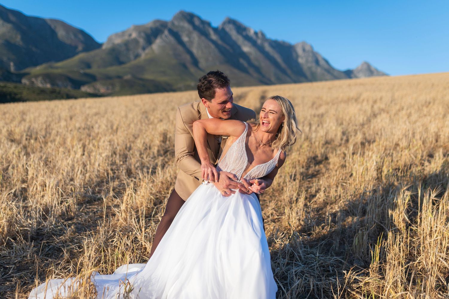 Couple in wedding attire laughing in golden wheat field with dramatic mountain backdrop on sunny day.
