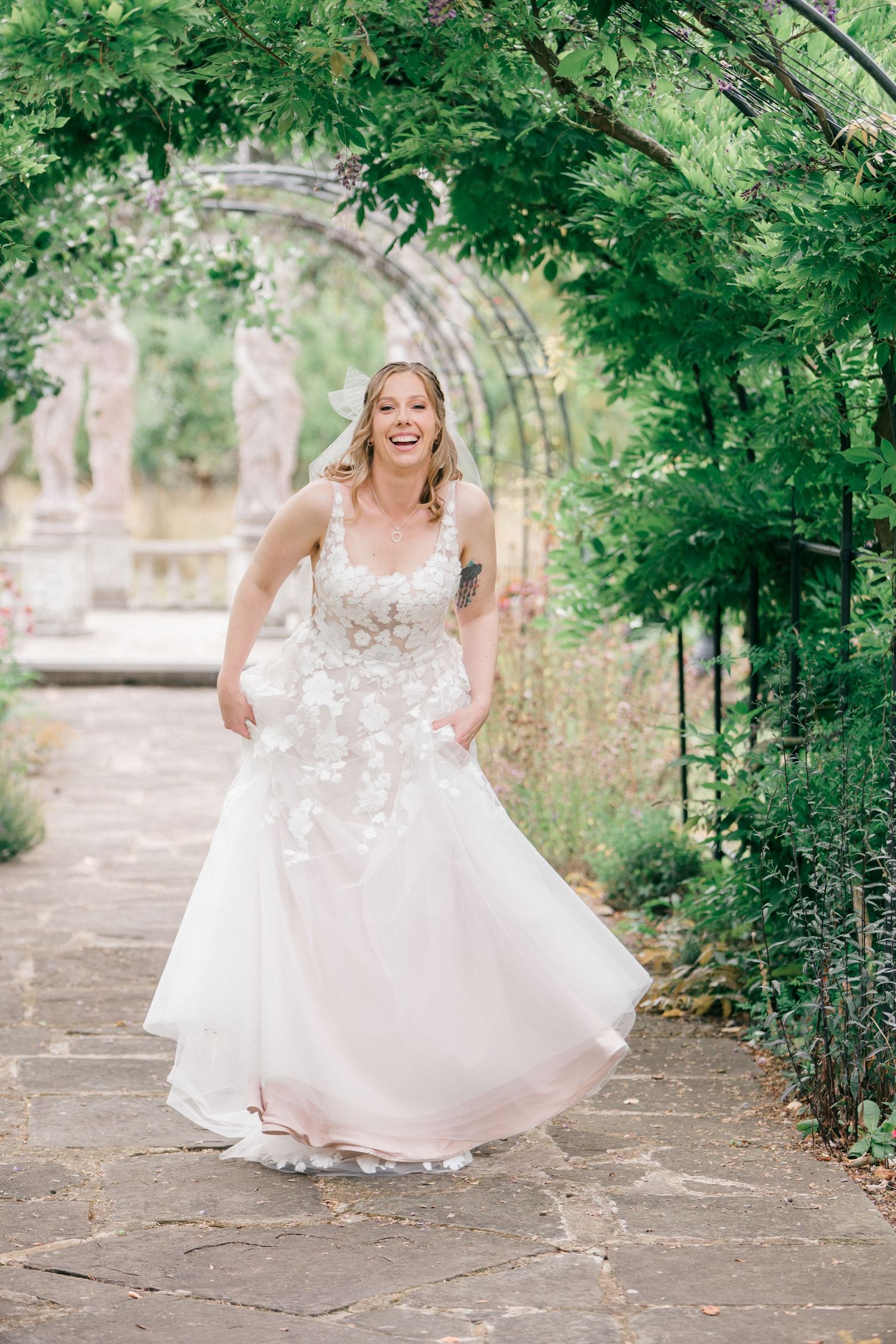 Bride in floral lace blush wedding gown laughing joyfully under green garden archway on stone path.
