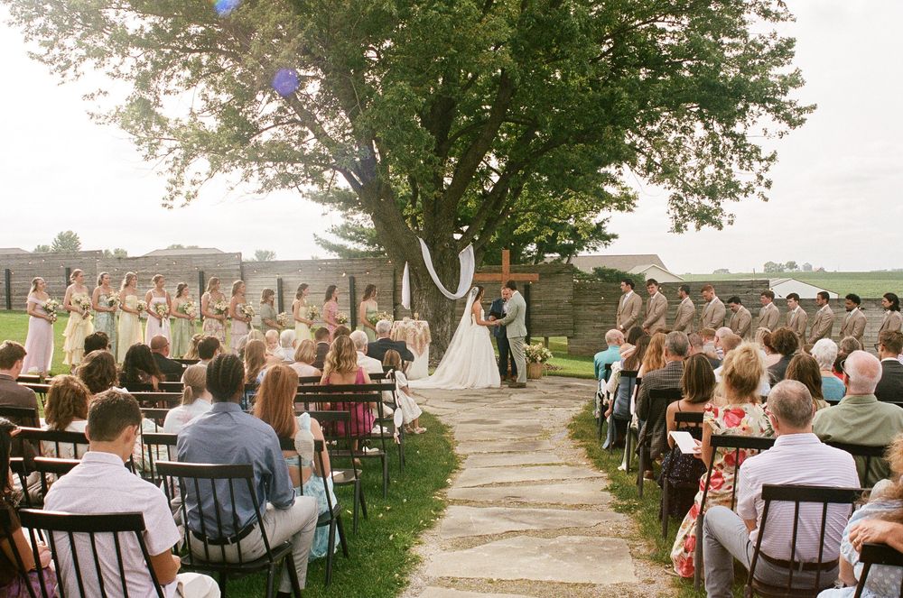 Outdoor wedding ceremony on stone path with bride and groom at altar, bridesmaids and groomsmen lined up under large tree.