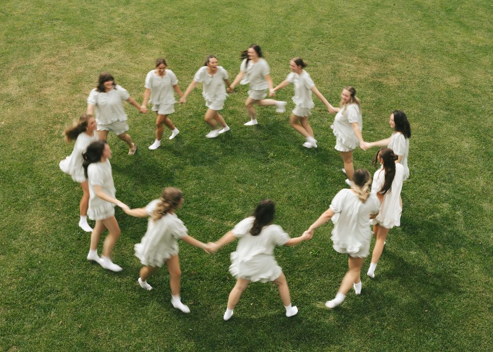 Girls in white dresses holding hands dancing in a circle on green grass outdoors.