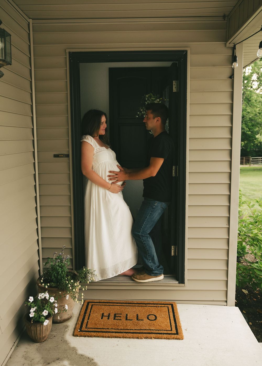 Pregnant woman in white dress with partner at front door on HELLO mat during maternity photo shoot.