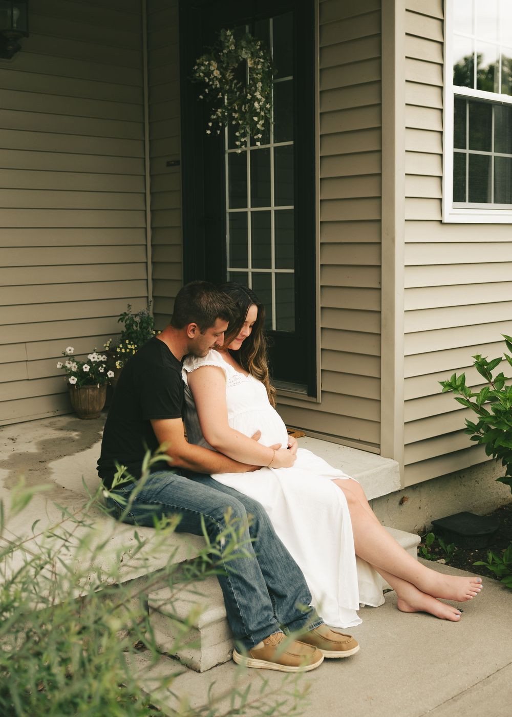 Couple sitting on front porch steps, man in black shirt and jeans embracing pregnant woman in white dress.
