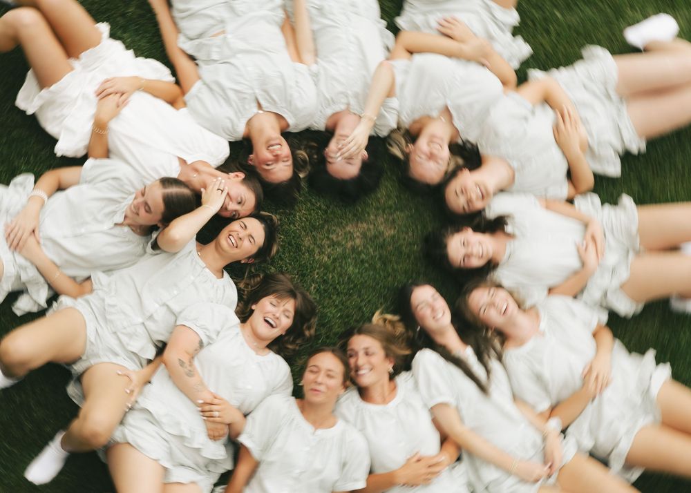 Group of women in white dresses lying in a circle on green grass, smiling and laughing, aerial view.