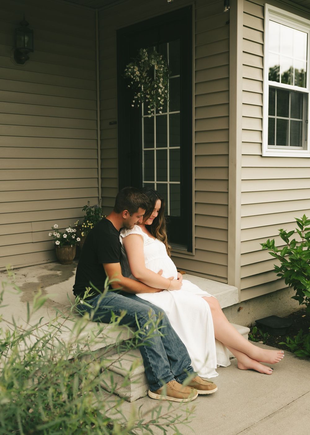 Couple sharing tender moment on porch steps, man kissing pregnant womans head in white dress.