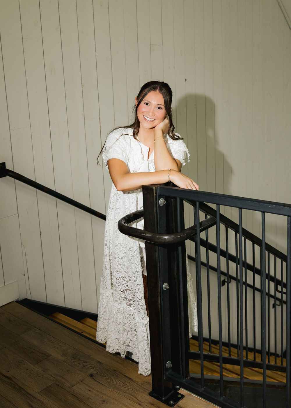 Young woman in white lace maxi dress posing gracefully on wooden staircase with black iron railing.