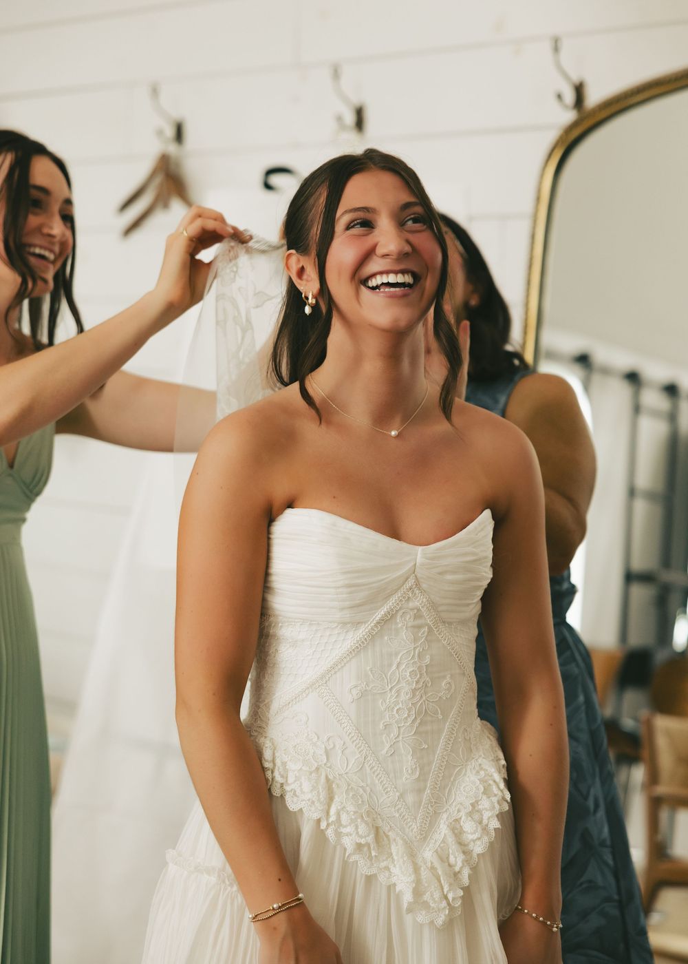 Bride laughing joyfully in a strapless white lace wedding dress while bridesmaids help adjust her veil.