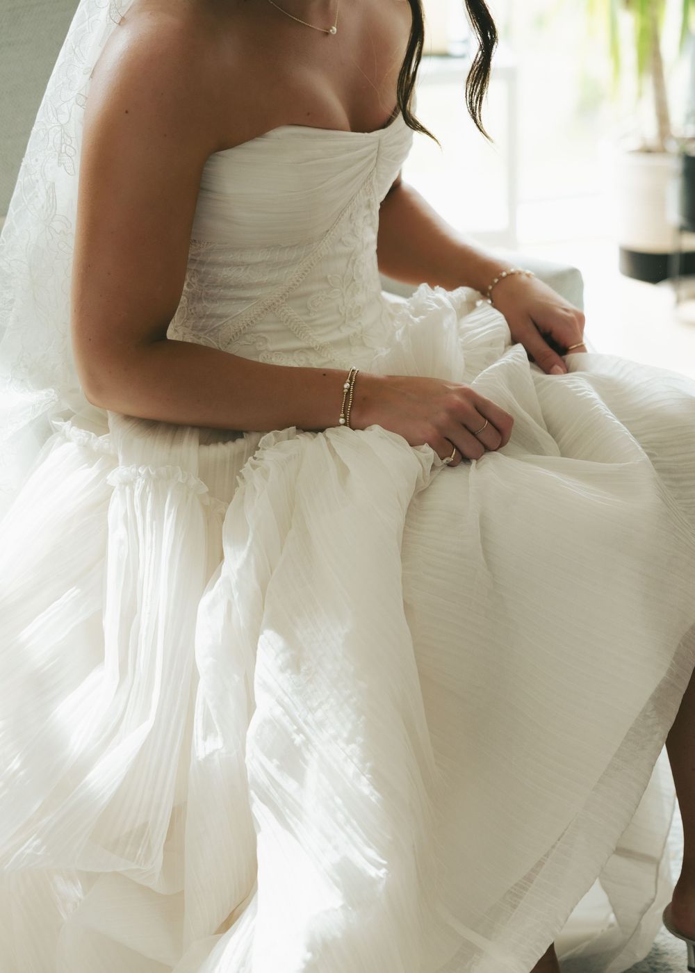 Bride in strapless white wedding gown with layered tulle skirt, wearing a delicate bracelet, seated elegantly.