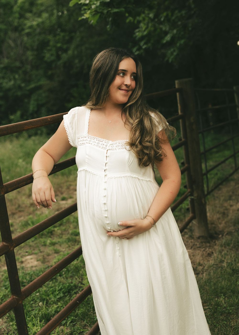Pregnant woman in white dress holding baby bump by rustic fence surrounded by lush greenery.