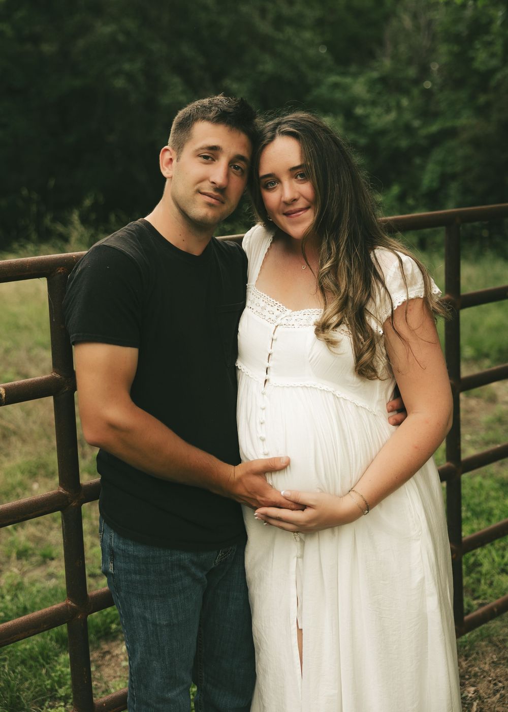 Couple posing outdoors near a rustic fence, woman in white dress cradling baby bump during maternity photo session.