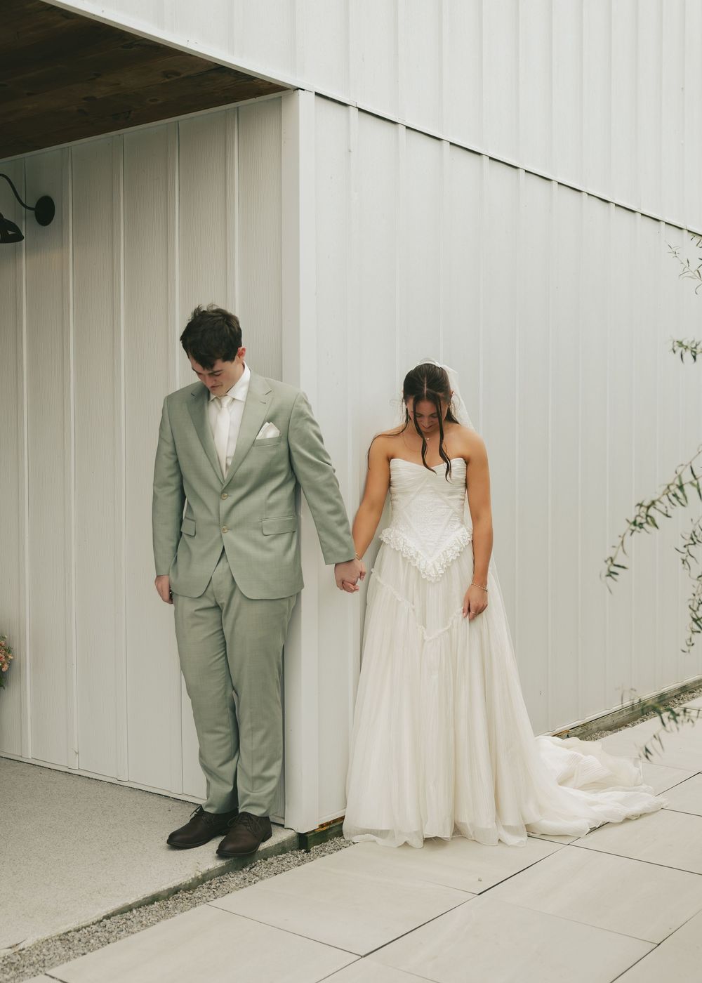 Bride and groom hold hands looking down outside a white barn venue in sage suit and strapless gown.