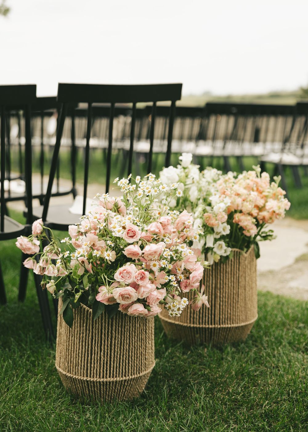 Woven seagrass baskets filled with blush pink roses and white flowers line a grass aisle at an outdoor wedding ceremony.