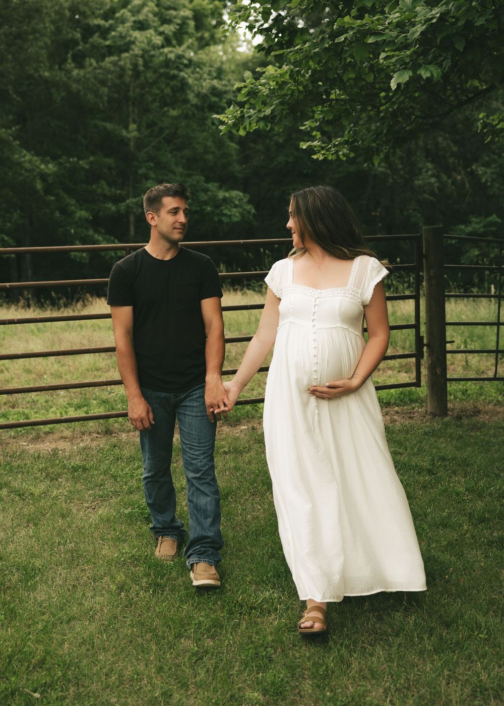 Pregnant woman in white dress holds hands with man in black shirt walking outdoors near rustic fence.
