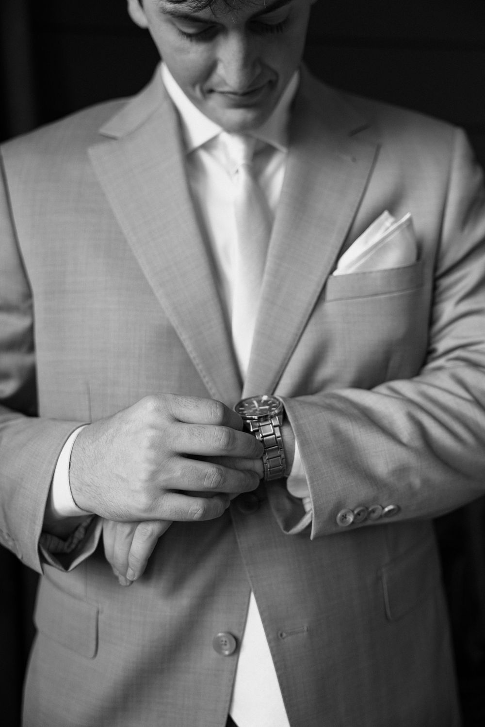 Groom in light gray suit adjusting his watch, with white pocket square, in black and white photo.