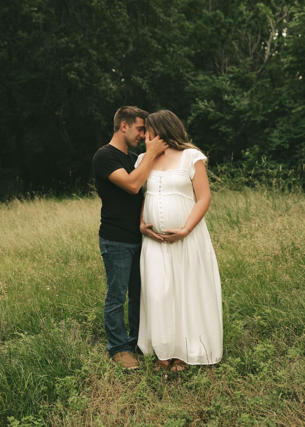 A couple shares a tender moment in a field, the woman in a white maternity dress cradling her baby bump.