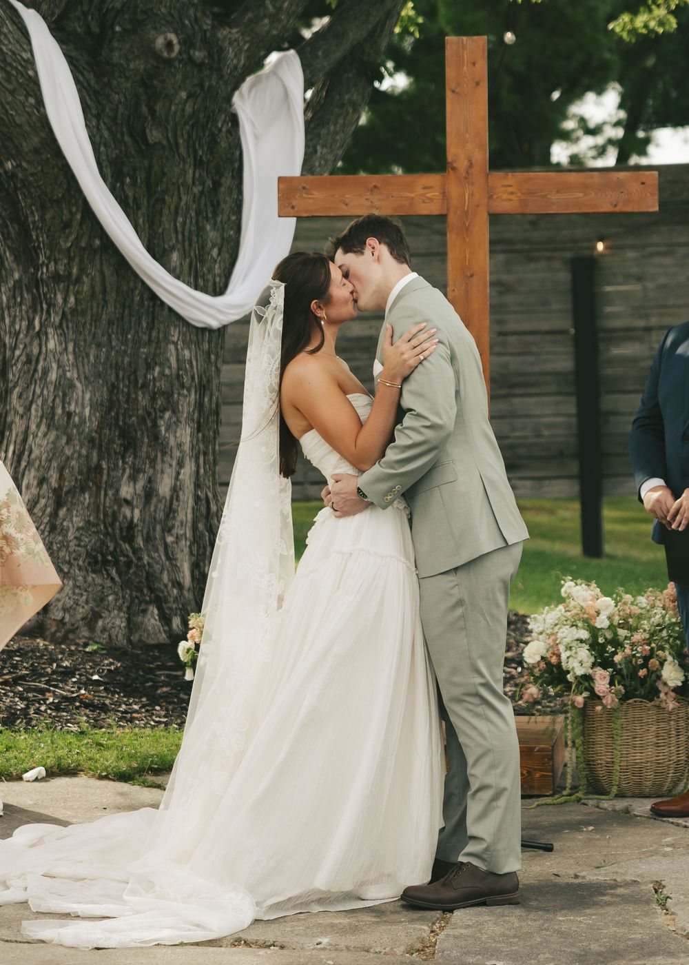 Bride and groom share first kiss at outdoor wedding ceremony beneath rustic wooden cross adorned with white fabric.