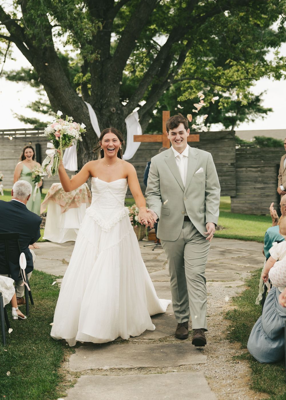 Joyful newlyweds walk down outdoor aisle after ceremony, bride in strapless gown holding bouquet, groom in sage suit.