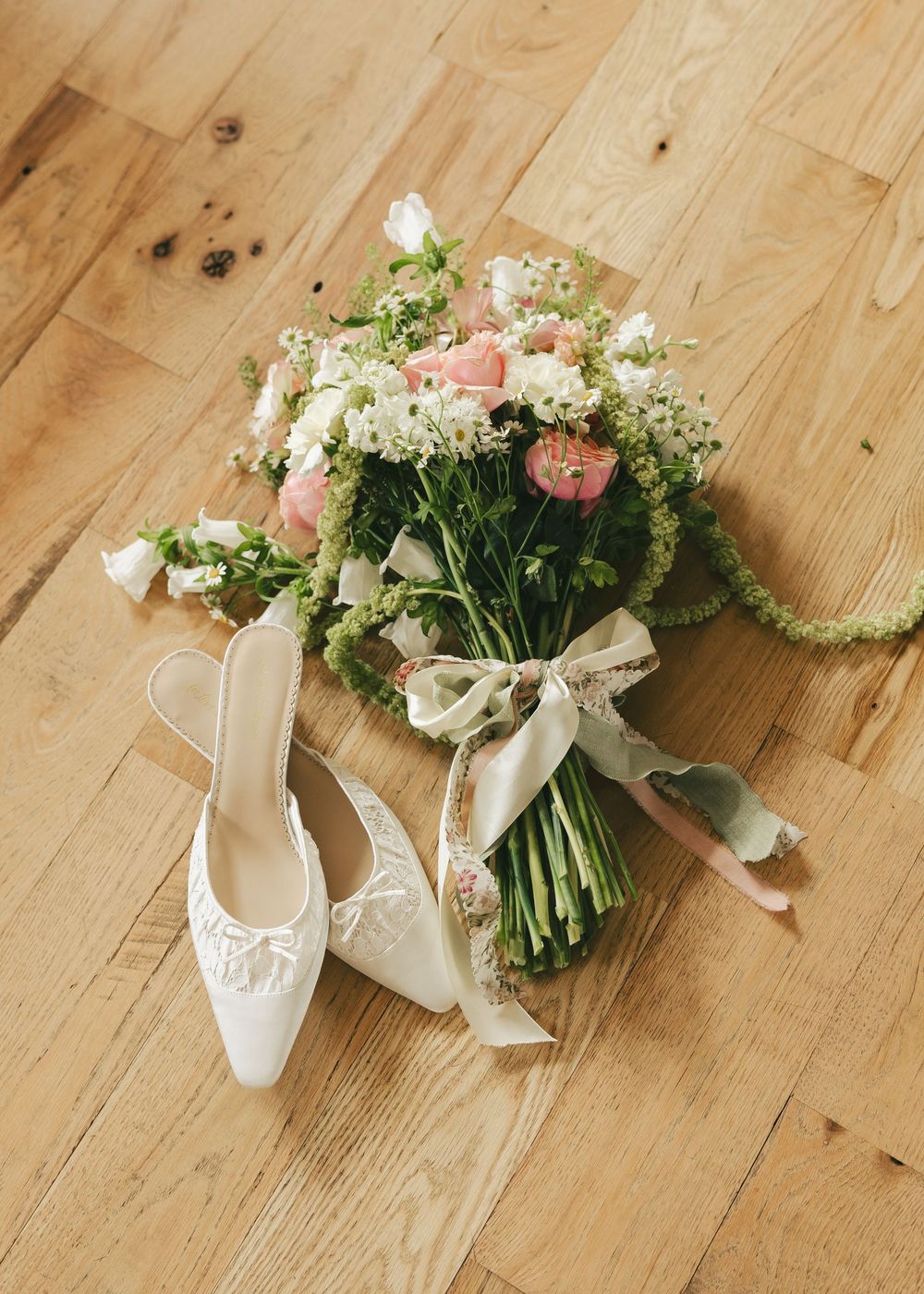 White bridal heels with lace detail beside a romantic pink and white wedding bouquet tied with ribbon on wood floor.