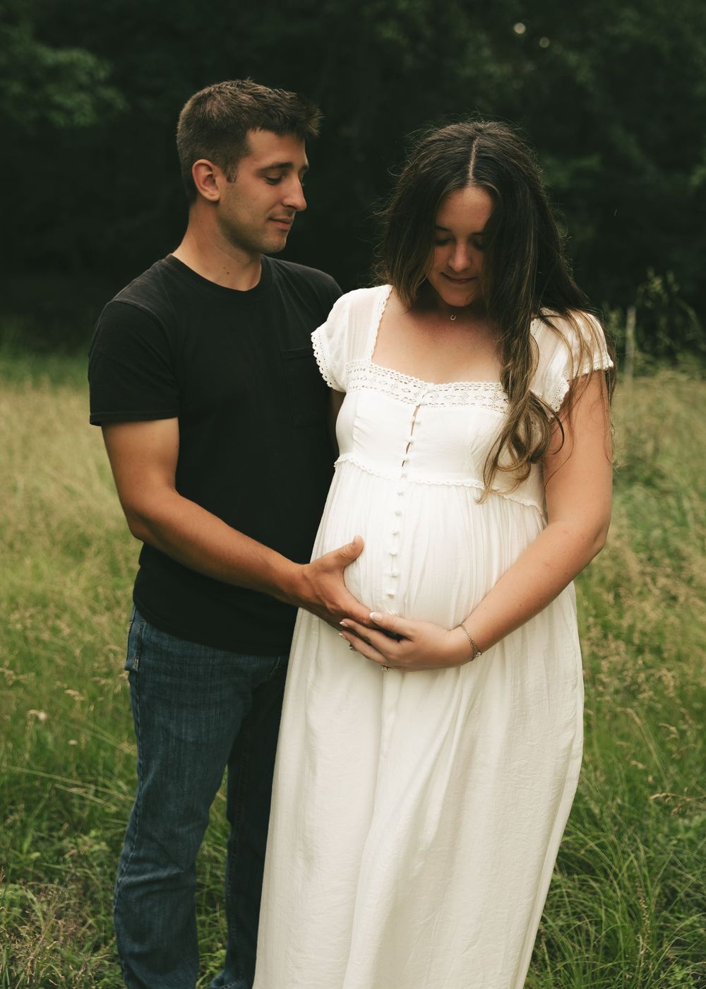 Couple posing outdoors in a field, man in black t-shirt embracing pregnant woman in white dress during maternity photoshoot.