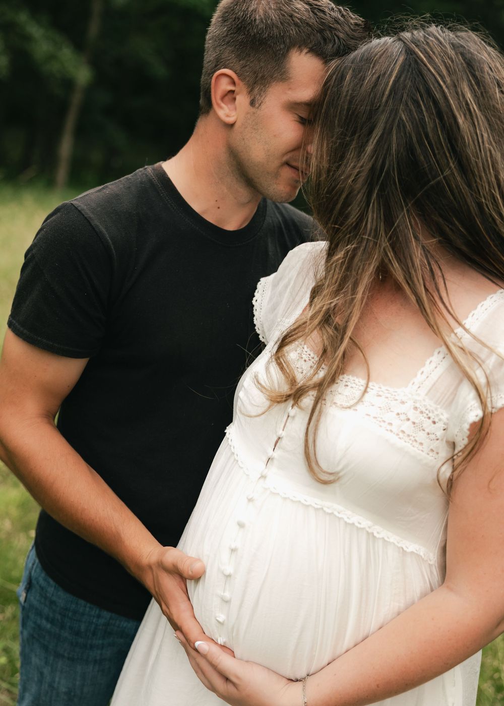 Couple sharing tender moment outdoors, man in black shirt kissing pregnant woman in white dress while cradling her baby bump.