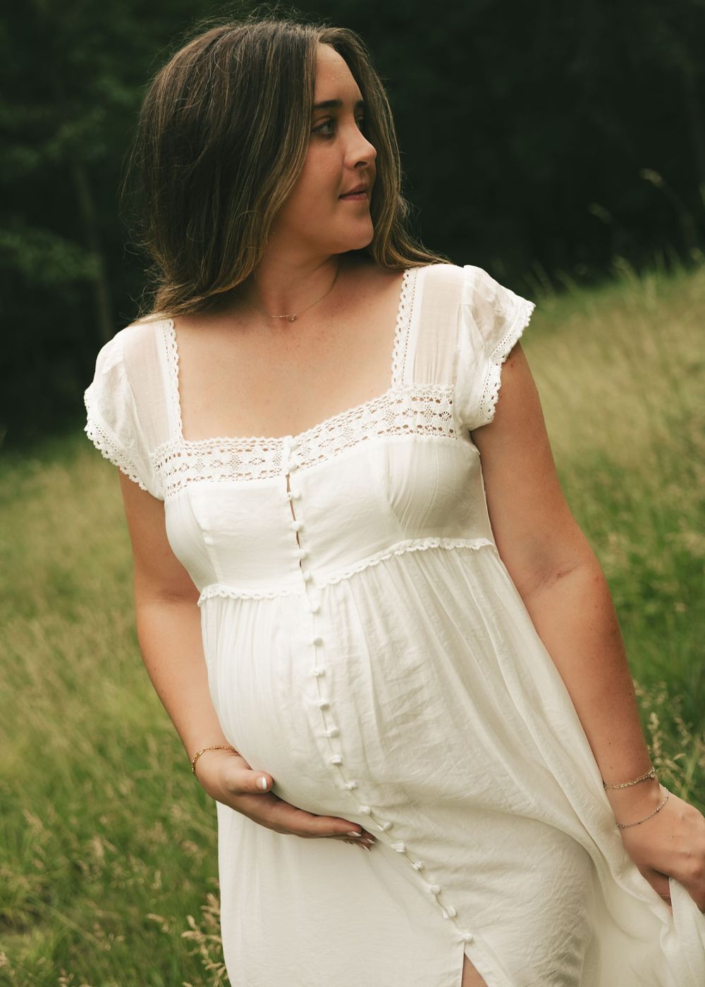 Pregnant woman in white lace dress cradling baby bump outdoors in a green field during maternity photoshoot.