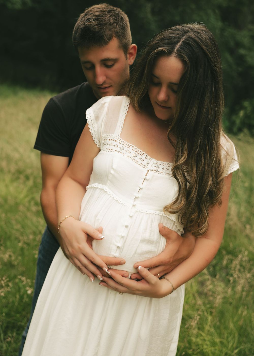 Couple in a field cradling pregnant belly, woman in white dress, maternity photo shoot outdoors.