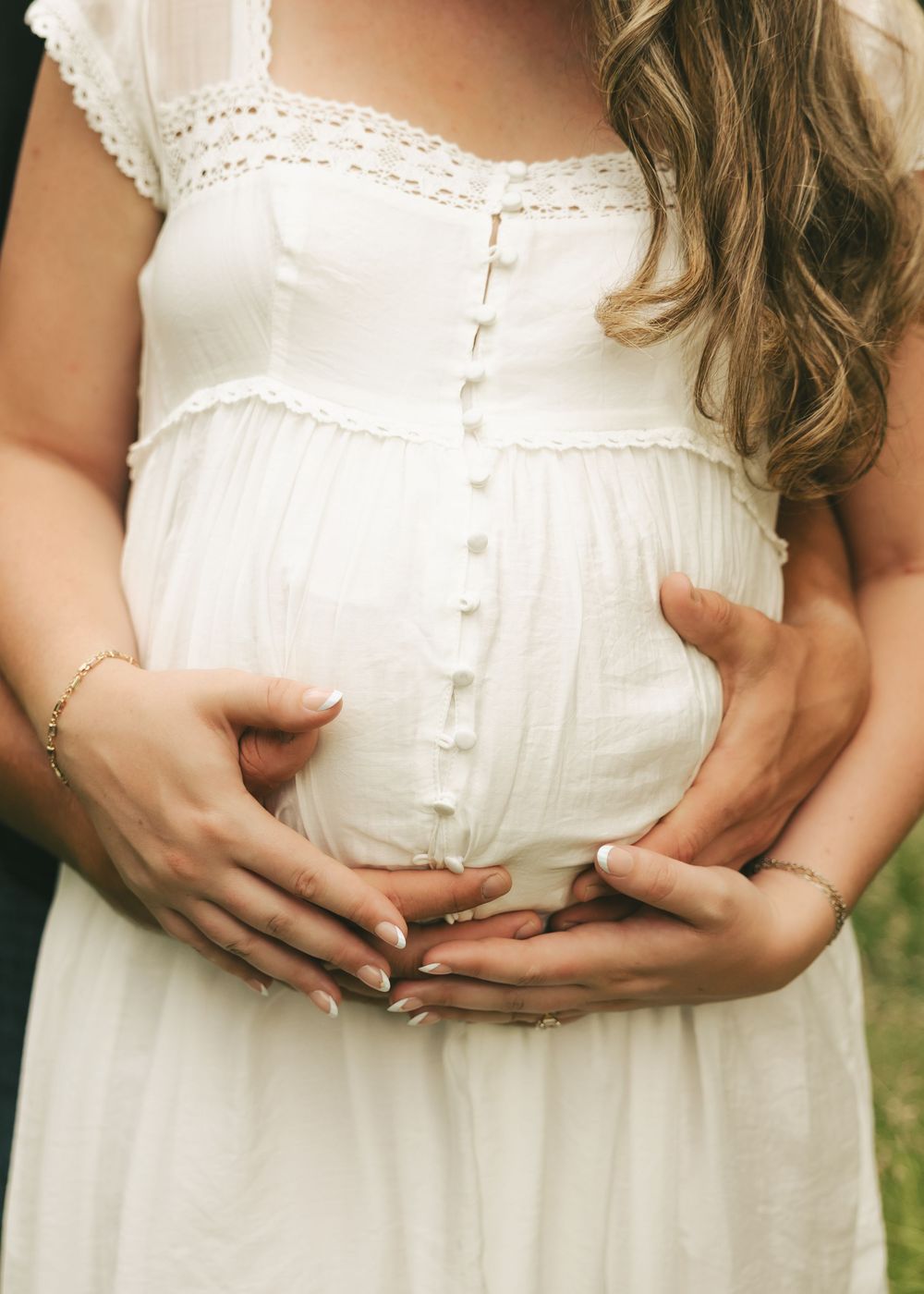 Close-up of pregnant woman in white button-down dress with couple's hands gently cradling her baby bump outdoors.