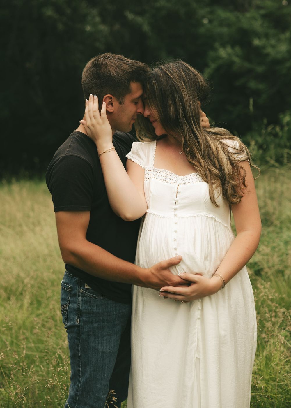 Couple sharing a tender moment in a field, man in black shirt embracing pregnant woman in white dress outdoors.