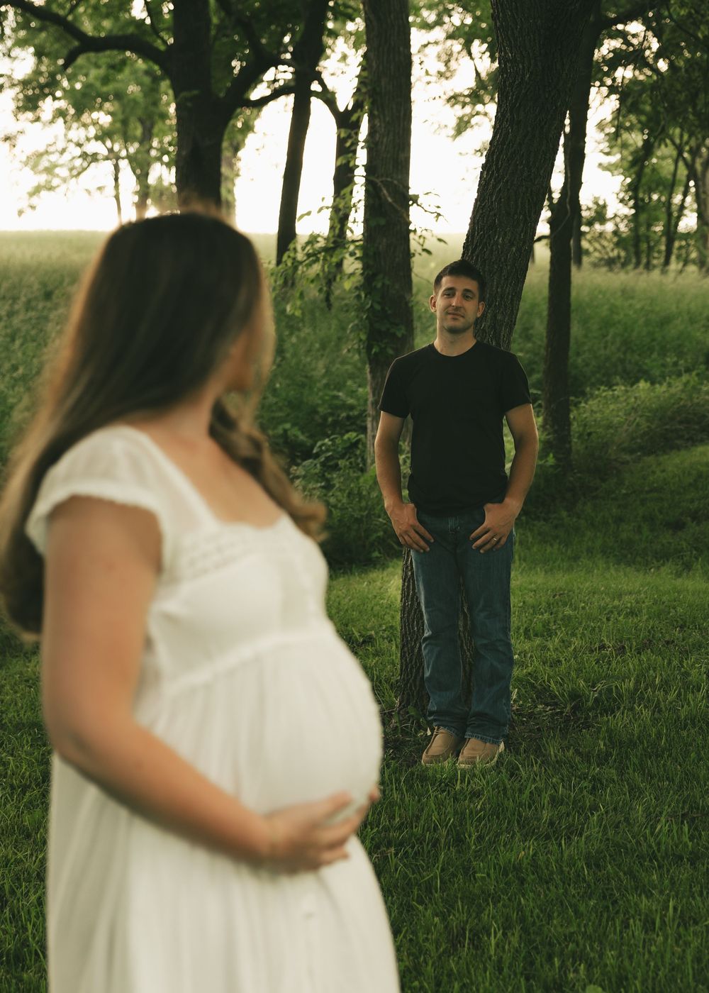Pregnant woman in white dress holds belly outdoors as man in black shirt stands by trees in soft evening light.