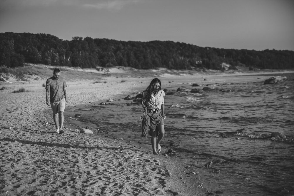 Black and white photo of a couple walking on a sandy beach near a stream with trees in the background.