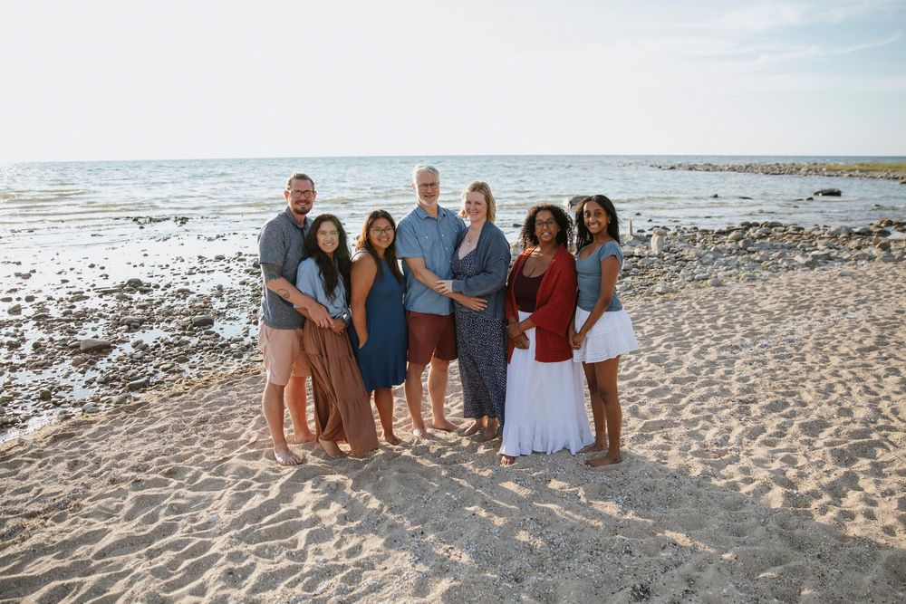 Family of seven smiling together on a sandy beach with ocean waves in the background on a sunny day.