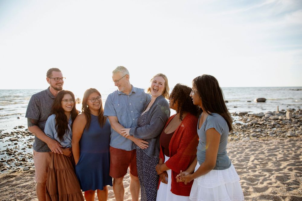 Group of seven friends smiling together on a sunny beach, casual attire, ocean backdrop, golden hour lighting.
