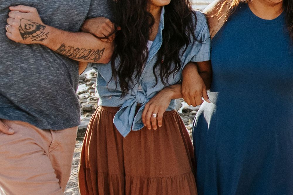 Three people standing close together outdoors, wearing blue and brown casual outfits, with a tattooed arm visible.