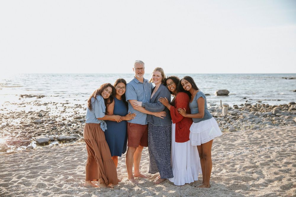 Happy family of six posing together on a sandy beach with ocean backdrop on a sunny day.