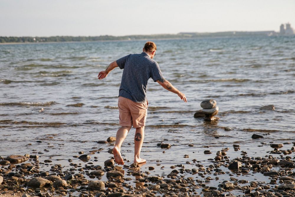 Man walking barefoot on rocky shoreline near calm ocean water on a sunny day.