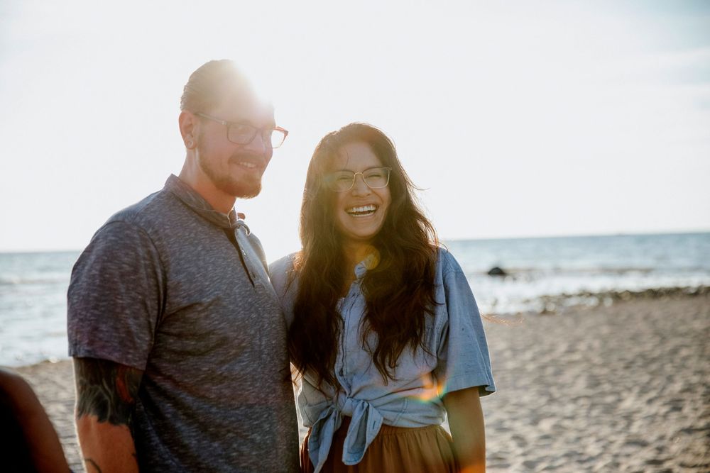 Couple smiling and laughing together on a sunny beach with ocean waves in the background.