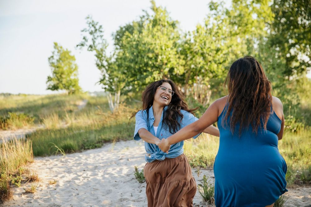 Two women laughing and holding hands on a sandy path surrounded by green trees on a sunny day.