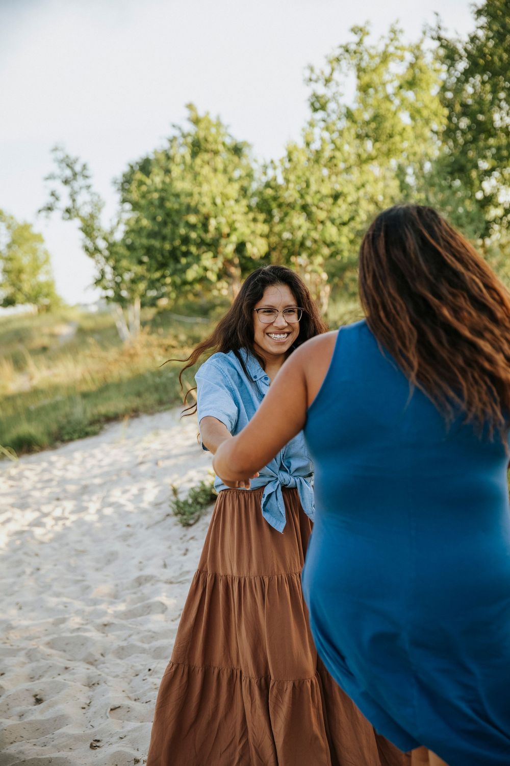 Two women holding hands on a sandy path, smiling joyfully, surrounded by green trees on a sunny day.