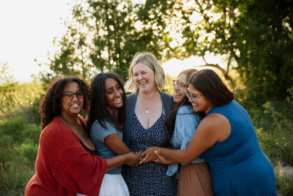 Five diverse women smiling and holding hands outdoors in golden hour light surrounded by lush green trees.
