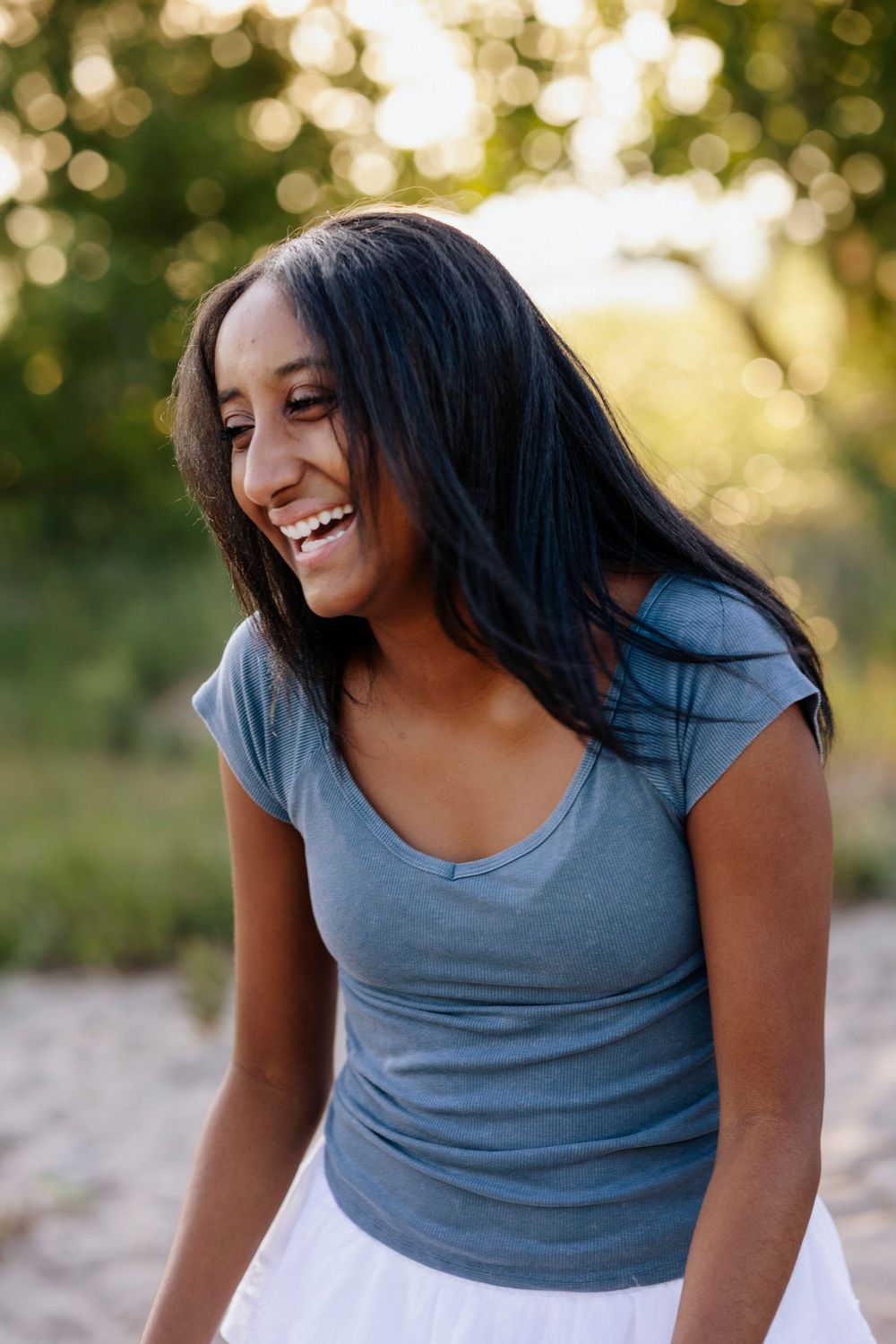 Young woman with long black hair laughing outdoors, wearing a blue t-shirt and white skirt in natural light.
