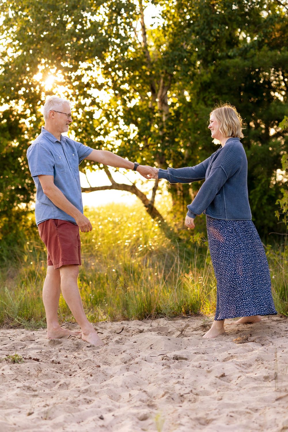 An older couple holds hands and dances barefoot on sandy beach at golden sunset with lush green trees behind them.