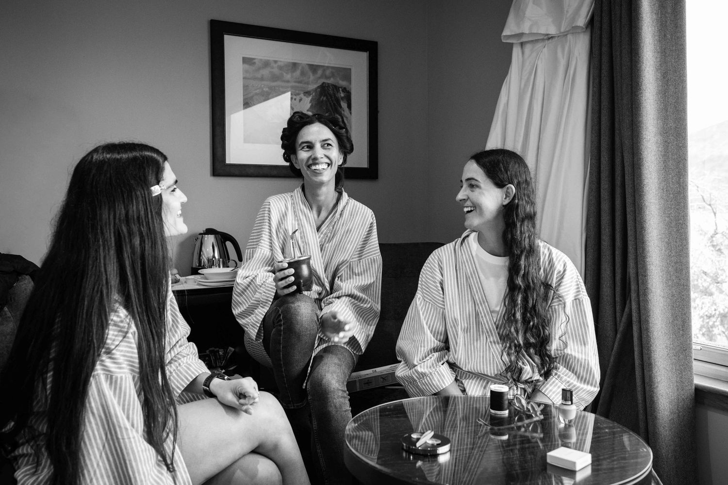 Three women laughing and chatting in a hotel room, one wearing a robe, in a candid black and white photo.
