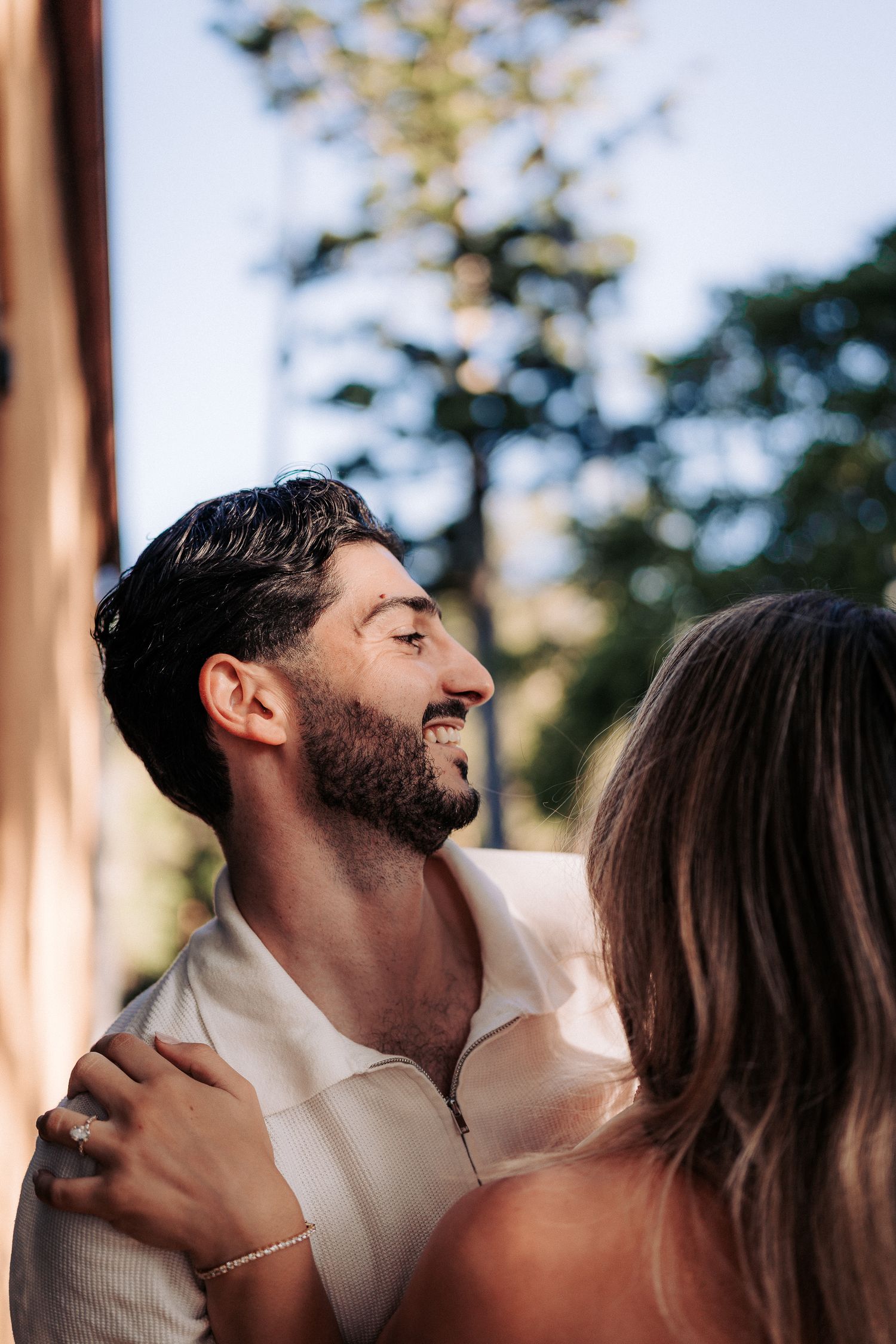 A smiling bearded man in a white shirt embraces a woman outdoors, with trees visible in the soft background.