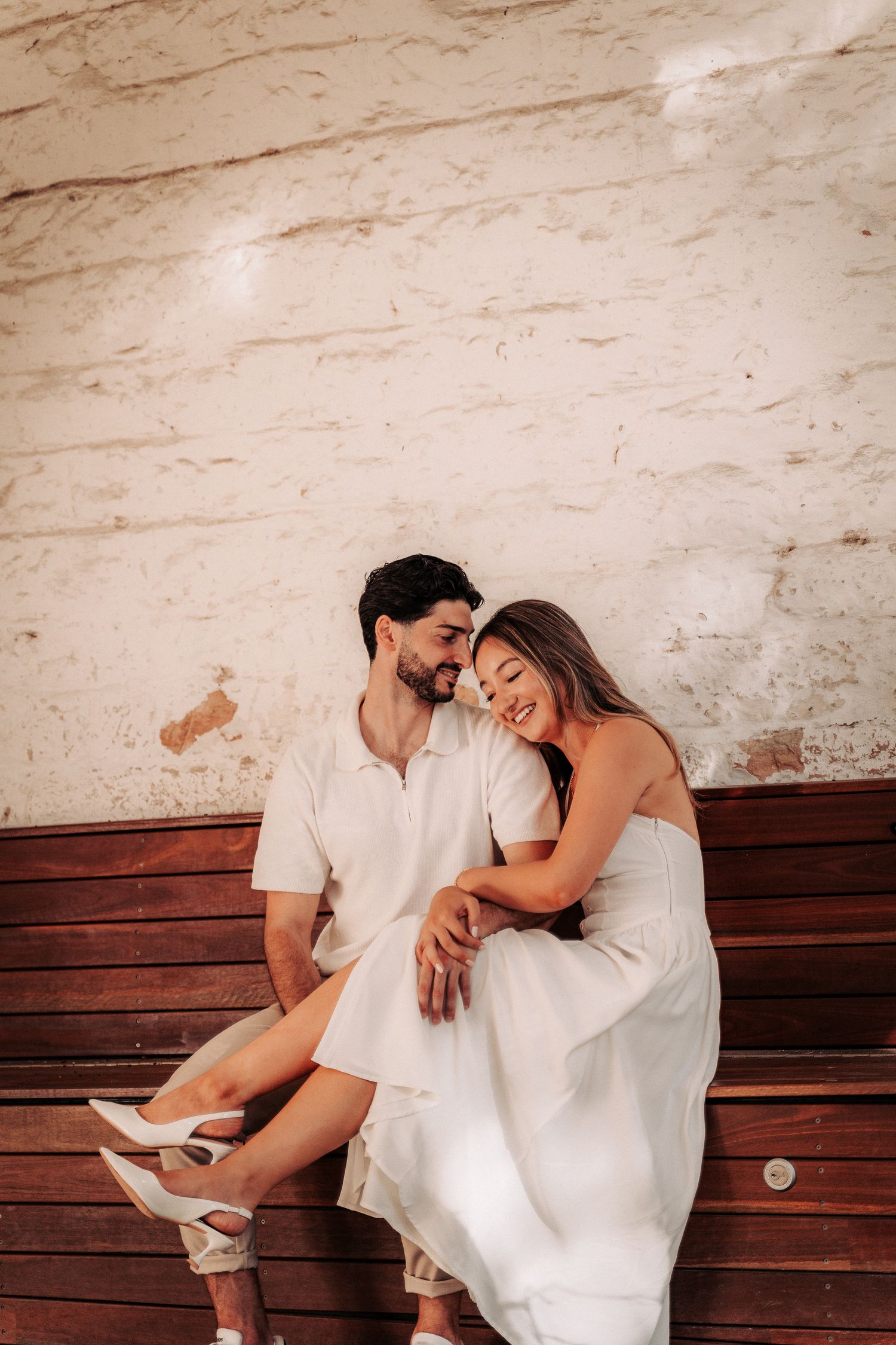 A couple in white outfits sitting closely on a wooden bench, smiling and embracing against a rustic wall.