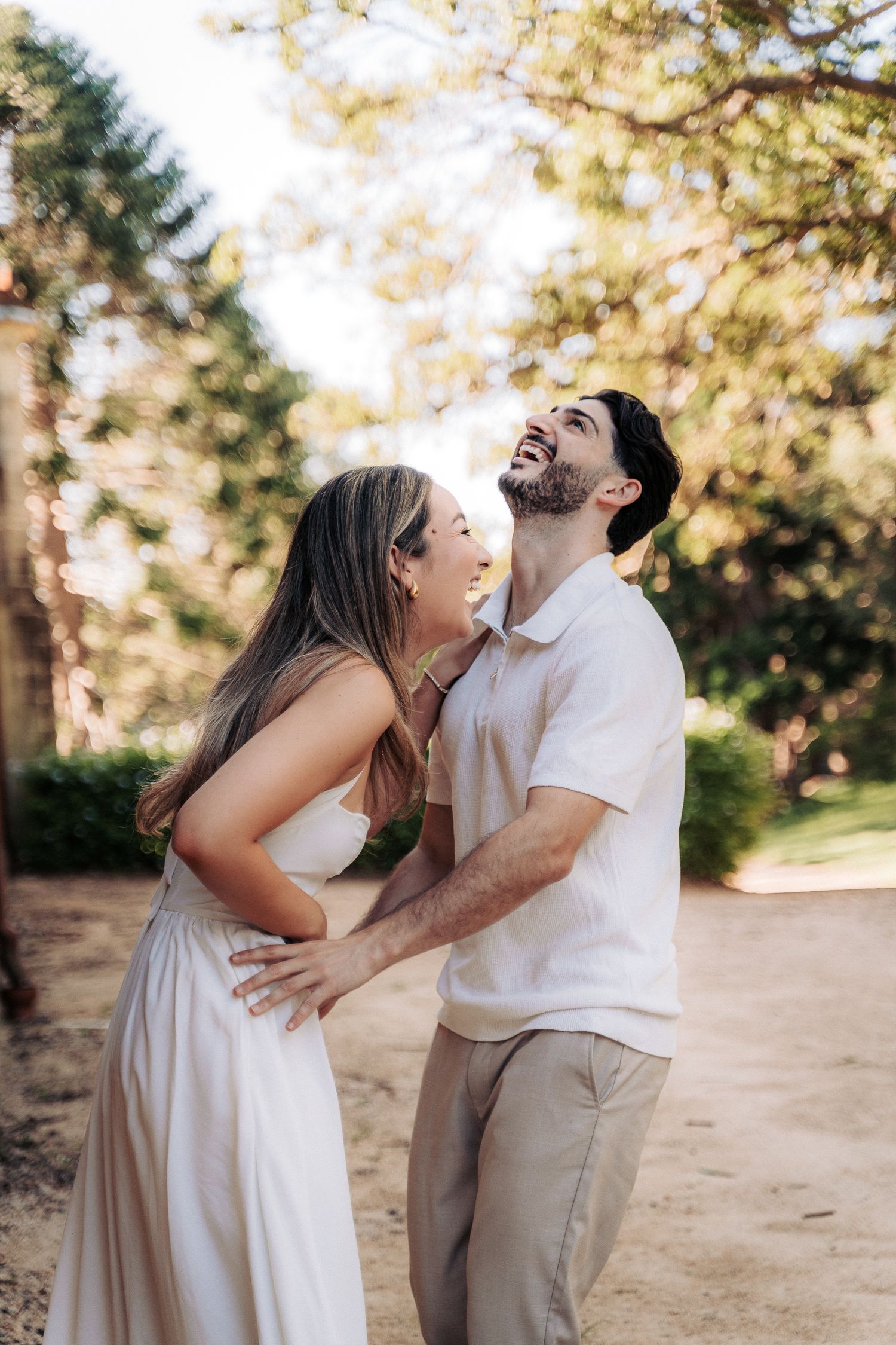 A joyful couple laughing together outdoors, woman in white dress embracing man in white polo shirt.